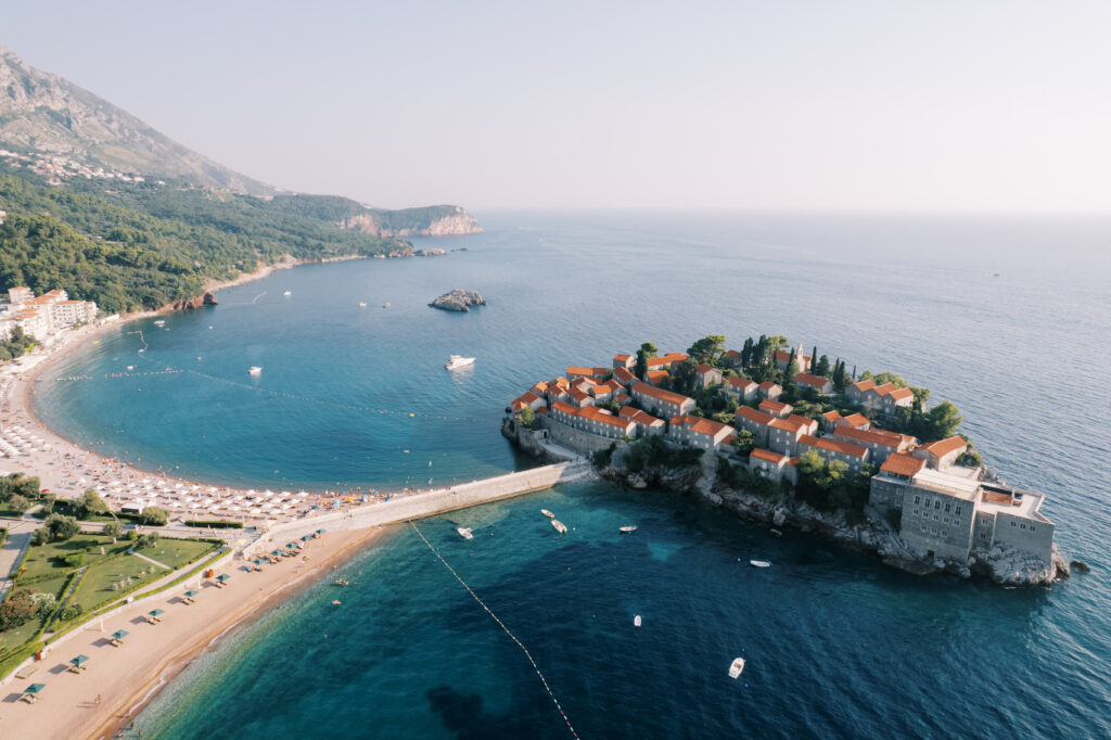 Vista aérea de la isla de Sveti Stefan, en Montenegro, que muestra el complejo hotelero Aman con sus edificios de tejados rojos rodeados por el mar Adriático. La isla está conectada a tierra firme por una estrecha pasarela. Playas de arena con sombrillas y bañistas bordean la costa. Hay barcos fondeados en las aguas azules y cristalinas. Al fondo se aprecian colinas y montañas verdes.